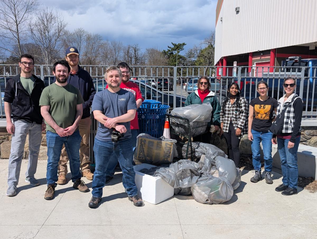 photo of a group of volunteers standing in front of large black trash bags and recyling bags with a blue sky with white clouds