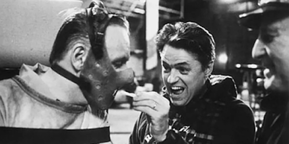 Actor Anthony Hopkins is wearing a straight jacket and restraint mask while director Jonathan Demme gives instructions.