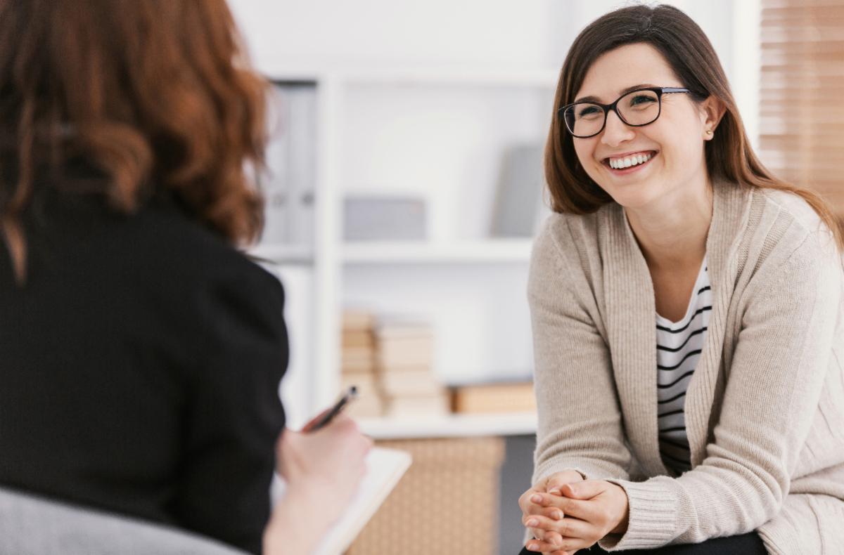Two people sitting indoors during a conversation; one person is holding a pen and notepad, the other is seated, smiling, with hands clasped, wearing a light cardigan and striped shirt.