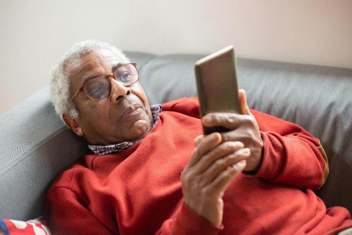 A person lying back on a couch, wearing a long‑sleeved red top, and holding a smartphone with both hands while looking at the screen.