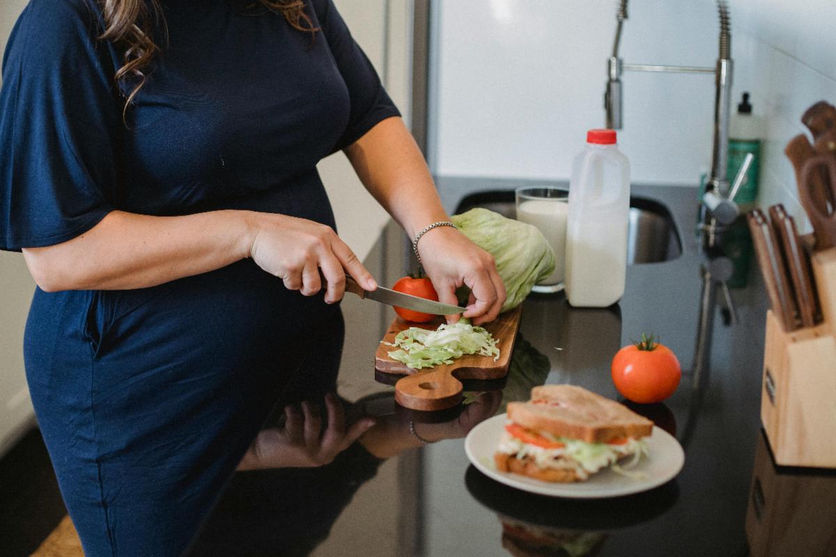 A pregnant person stands at a kitchen counter slicing a tomato on a cutting board with lettuce. A prepared sandwich sits on a plate nearby, along with fresh ingredients like a tomato and milk, suggesting healthy meal preparation at home.