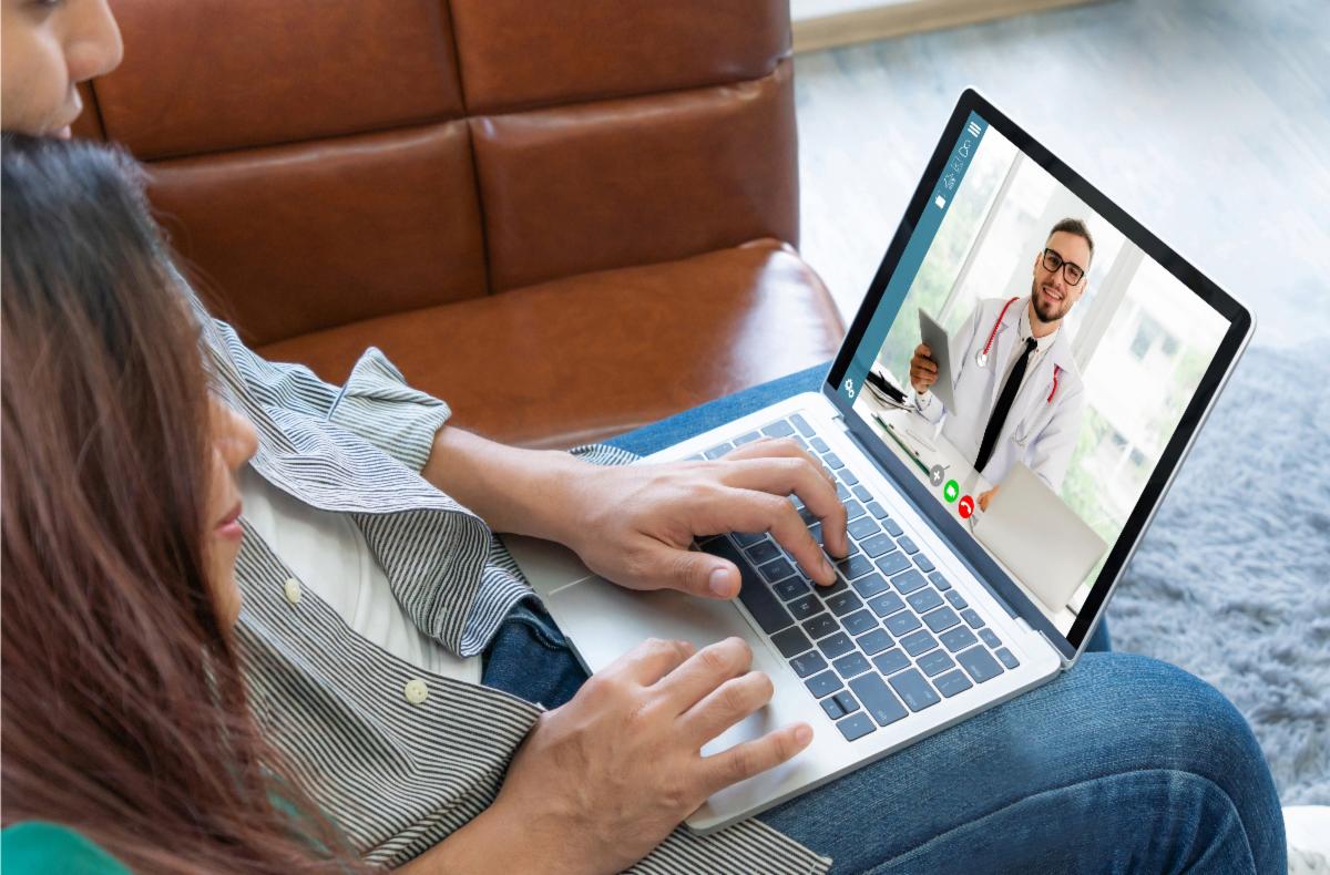 Banner image: A man and a woman sitting on a couch using a laptop for a telehealth video call with a doctor, who is shown on the screen wearing a white coat and stethoscope.