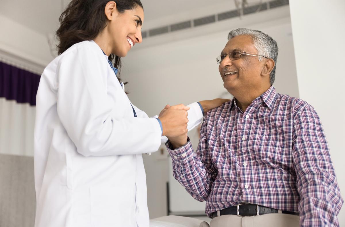Healthcare professional in a white coat holding the hand of a seated person wearing a checkered shirt, in a clinical setting.