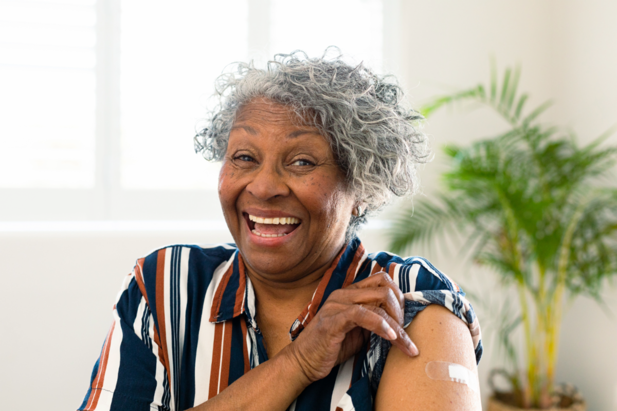 Older woman smiling, pointing to bandage on upper arm, post vaccination.