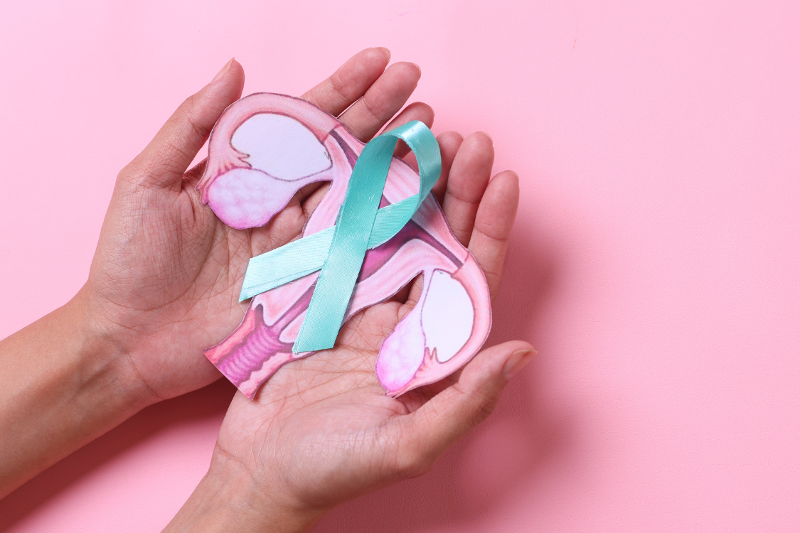 Two hands hold a paper cutout of the female reproductive system with a teal awareness ribbon placed across it, set against a pink background. The image symbolizes awareness and support for ovarian cancer and polycystic ovary syndrome (PCOS).