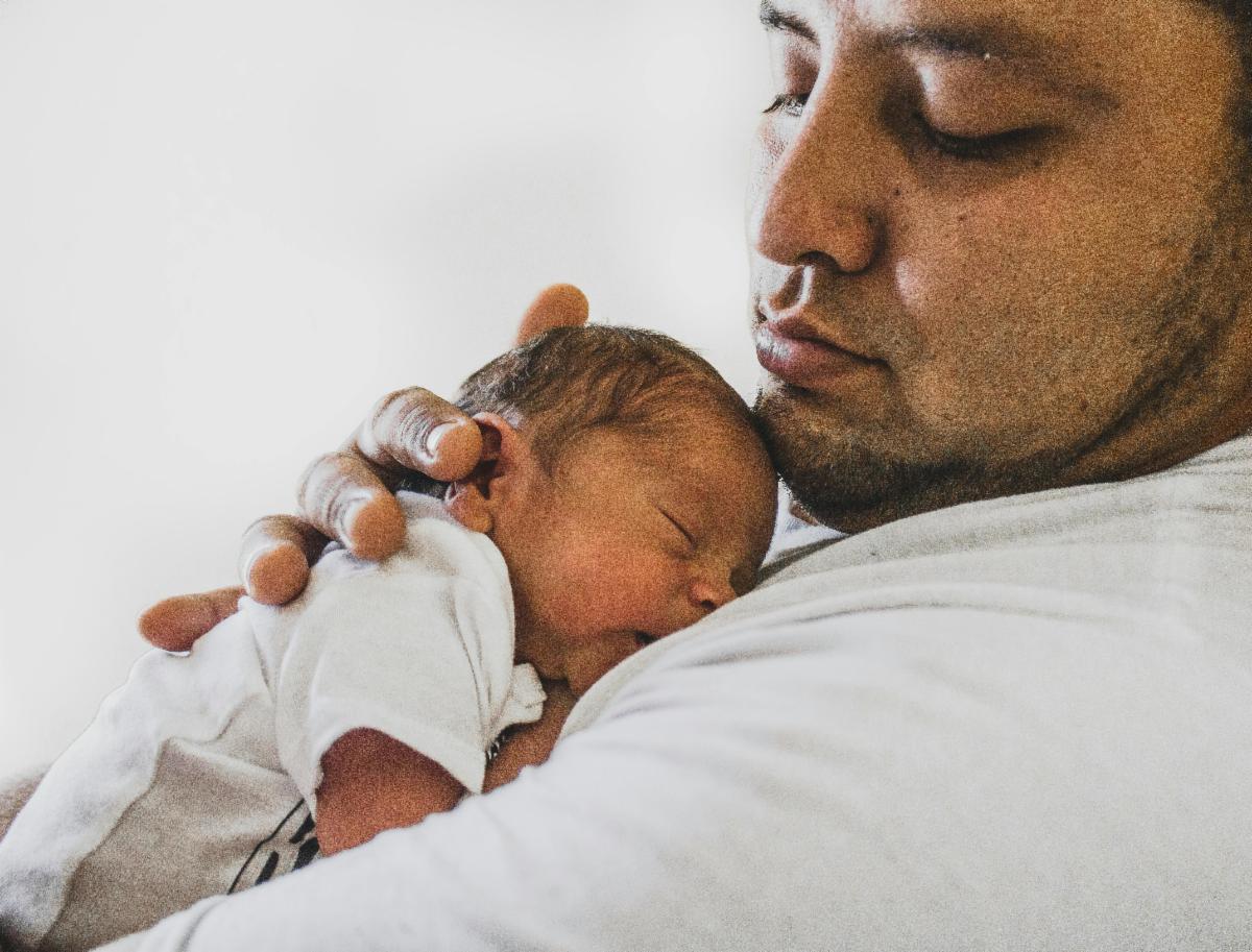 An adult cradles a sleeping newborn against their chest, gently supporting the baby’s head with one hand. The baby is dressed in a light-colored outfit, and the close-up composition emphasizes warmth, care, and bonding.
