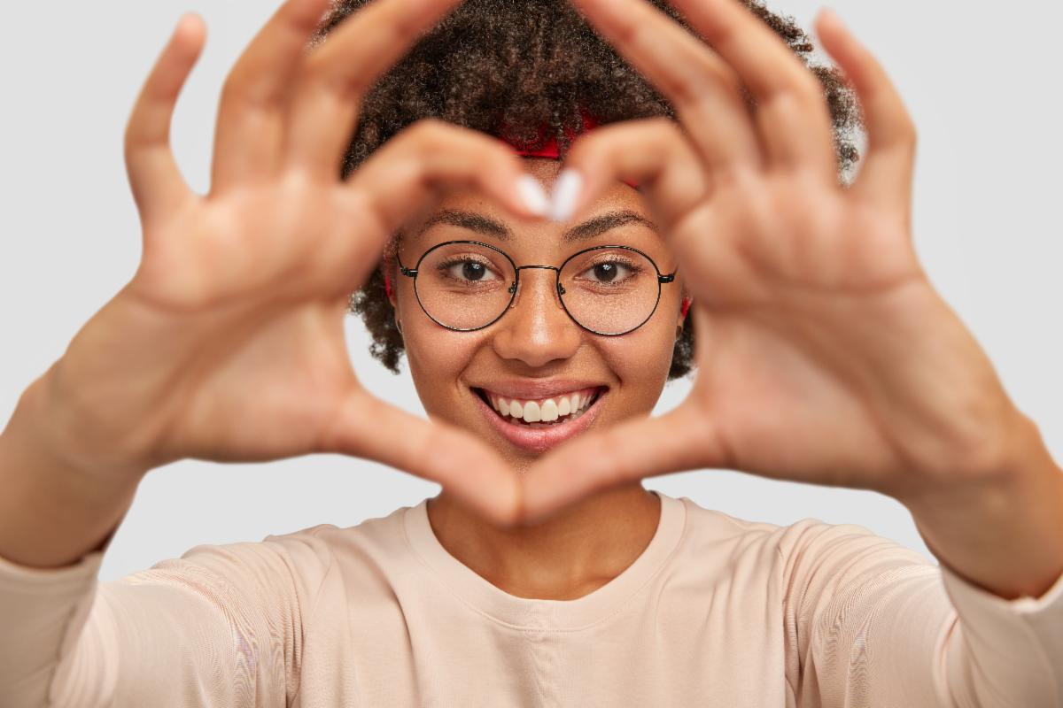 A person holding both hands up close to the camera, forming a circular or heart like shape with their fingers. The individual is wearing a long sleeved light-colored top, and their head is positioned behind their hands.