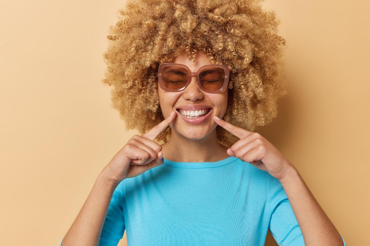 Person pointing to the sides of the mouth while wearing a light blue shirt against a tan background.