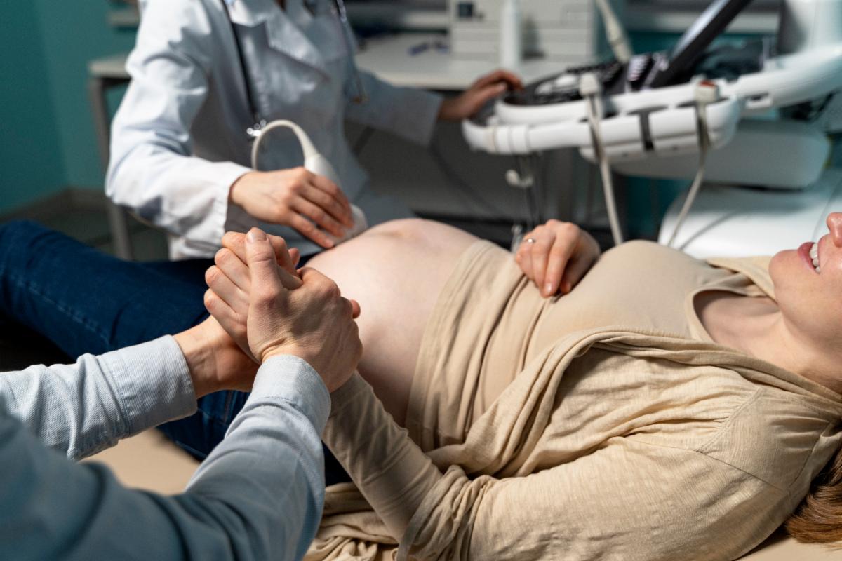 A pregnant person lies on an exam table during a prenatal ultrasound while a clinician uses an ultrasound probe on the abdomen. Another adult sits nearby holding the pregnant person’s hand for support, and medical equipment and monitors are visible in the clinical setting.