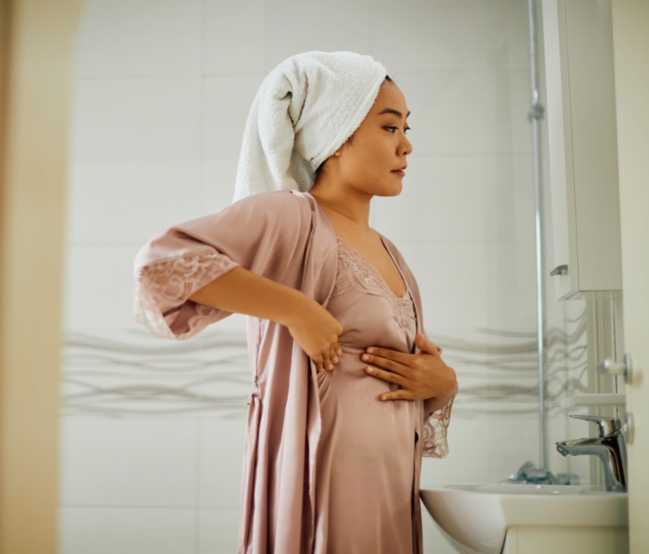 a woman wearing a towl on her head and a silk-like nightgown and robe examines the side of her breast with her hands. Related to breast cancer awareness month.