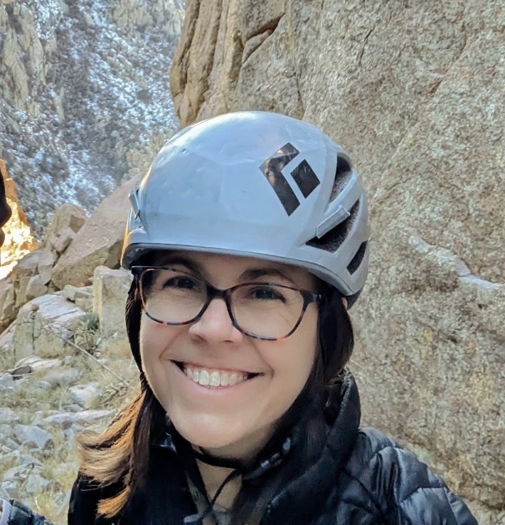 Woman wearing a gray rock climbing helmet smiling with a granite crag behind her