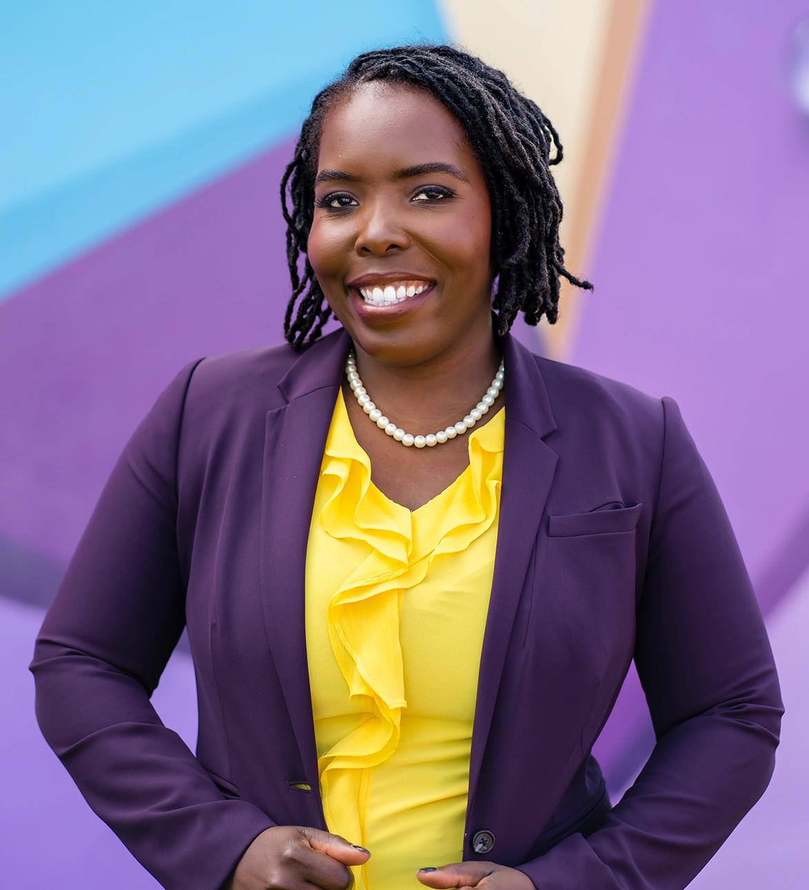 Flo Cofer - a Black woman wearing a purple jacket and yellow blouse.