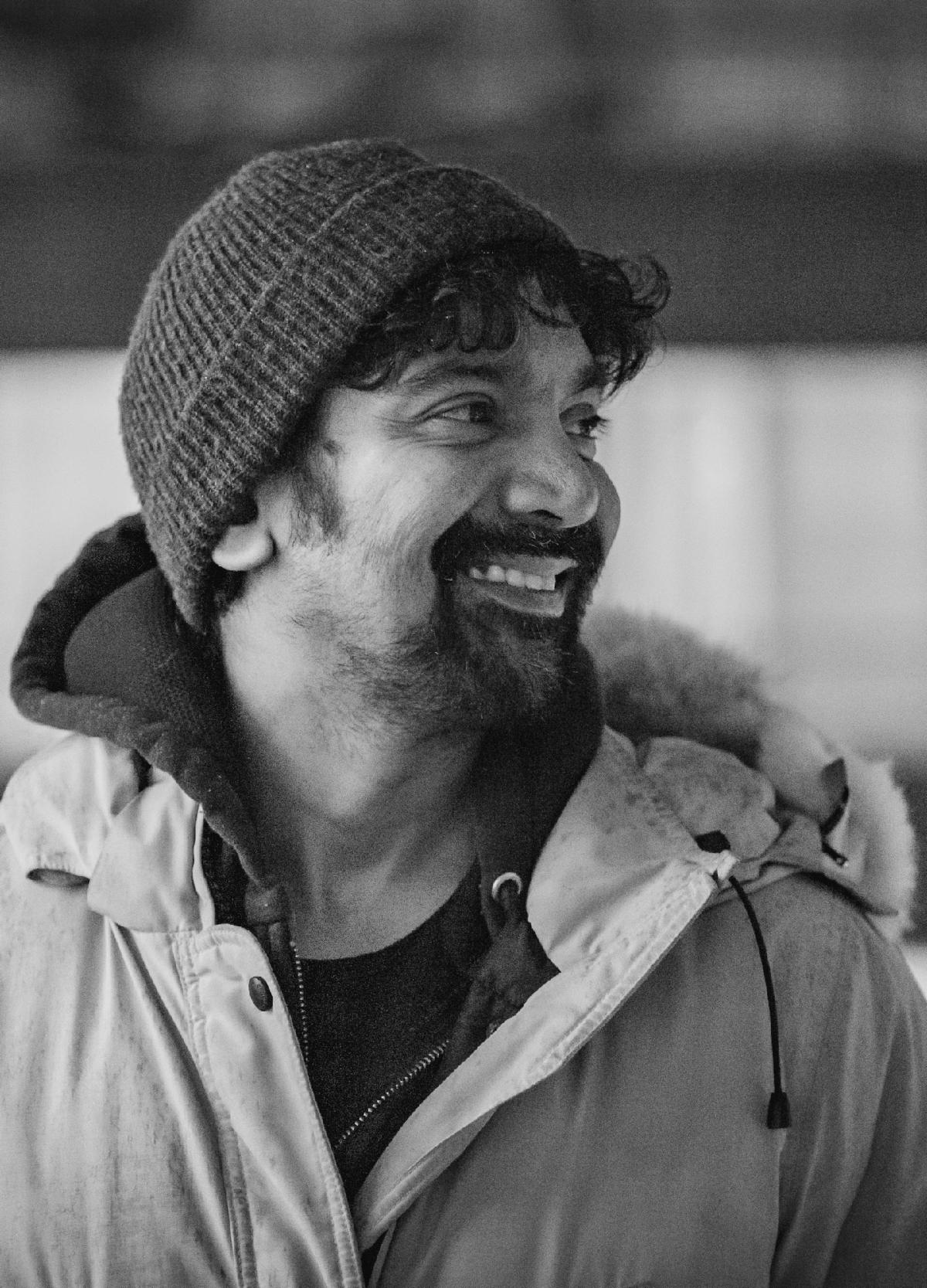 Black and white image of Hari Alluri in front of a blurred apartment building. Hari is bearded and smiling while facing the right of camera. Hari has black hair tufting out the front and wears a winter jacket with a black ribbed toque.
