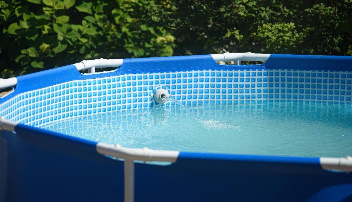 A blue above-ground swimming pool filled with clear water sits outdoors surrounded by green trees.