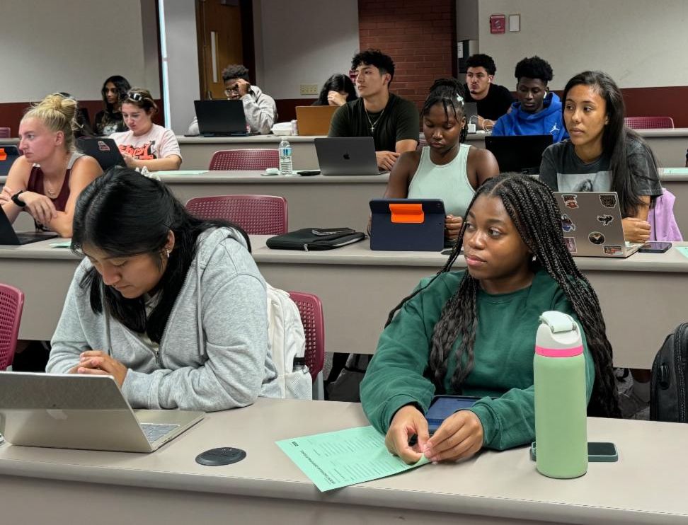 Student in classroom pointing to whiteboard while writing on paper with blue marker