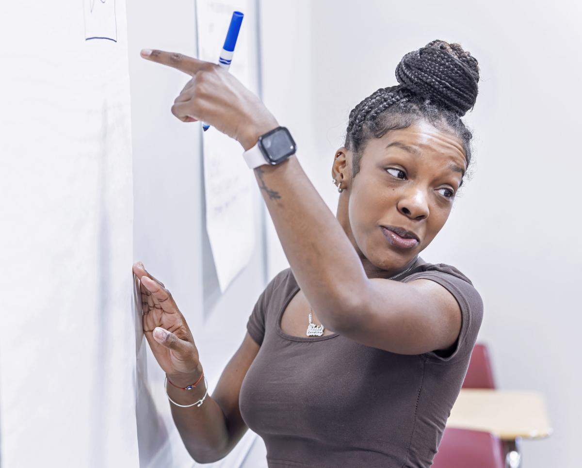 Student in classroom pointing to whiteboard while writing on paper with blue marker