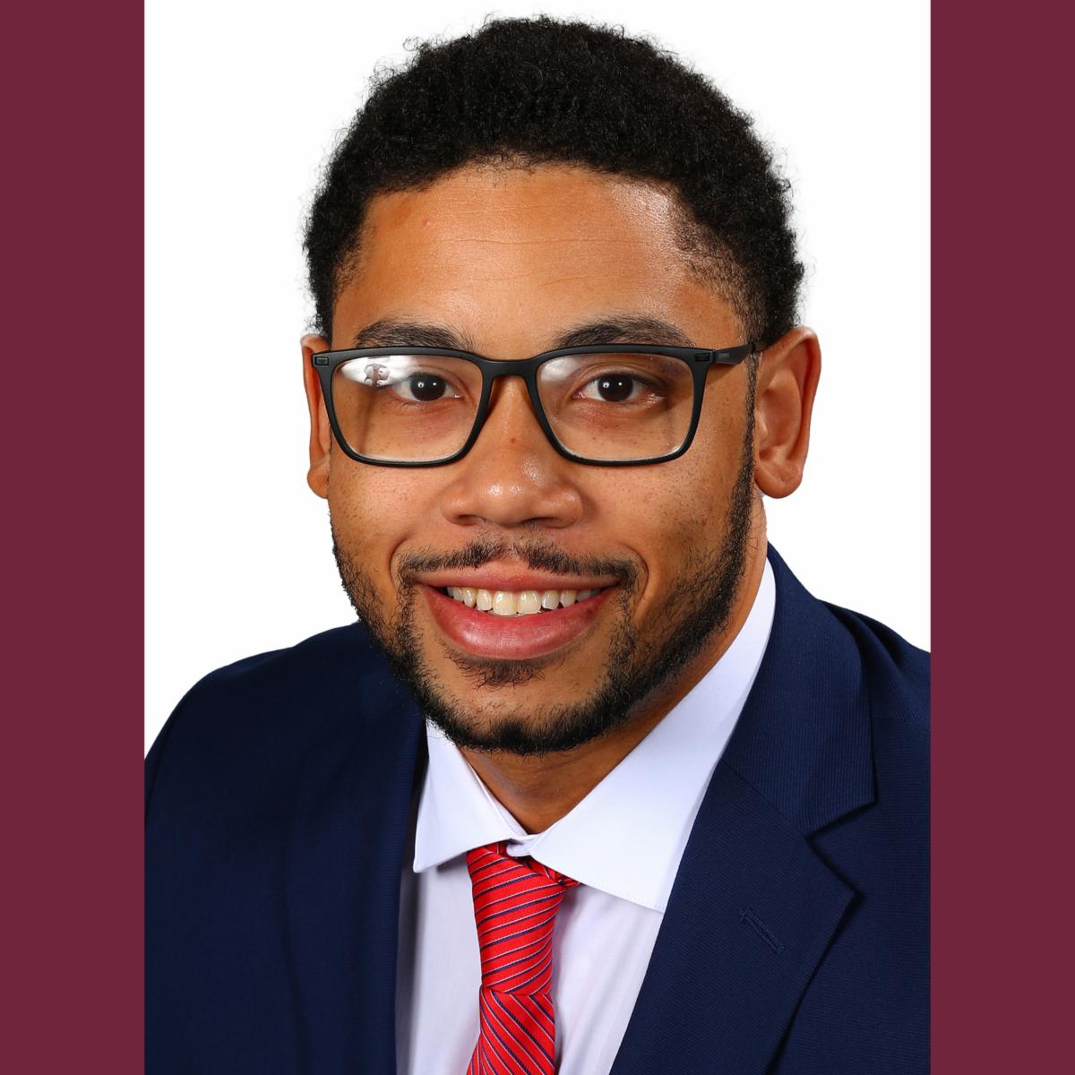 Kameron smiling wearing suit and tie and black rimmed glasses