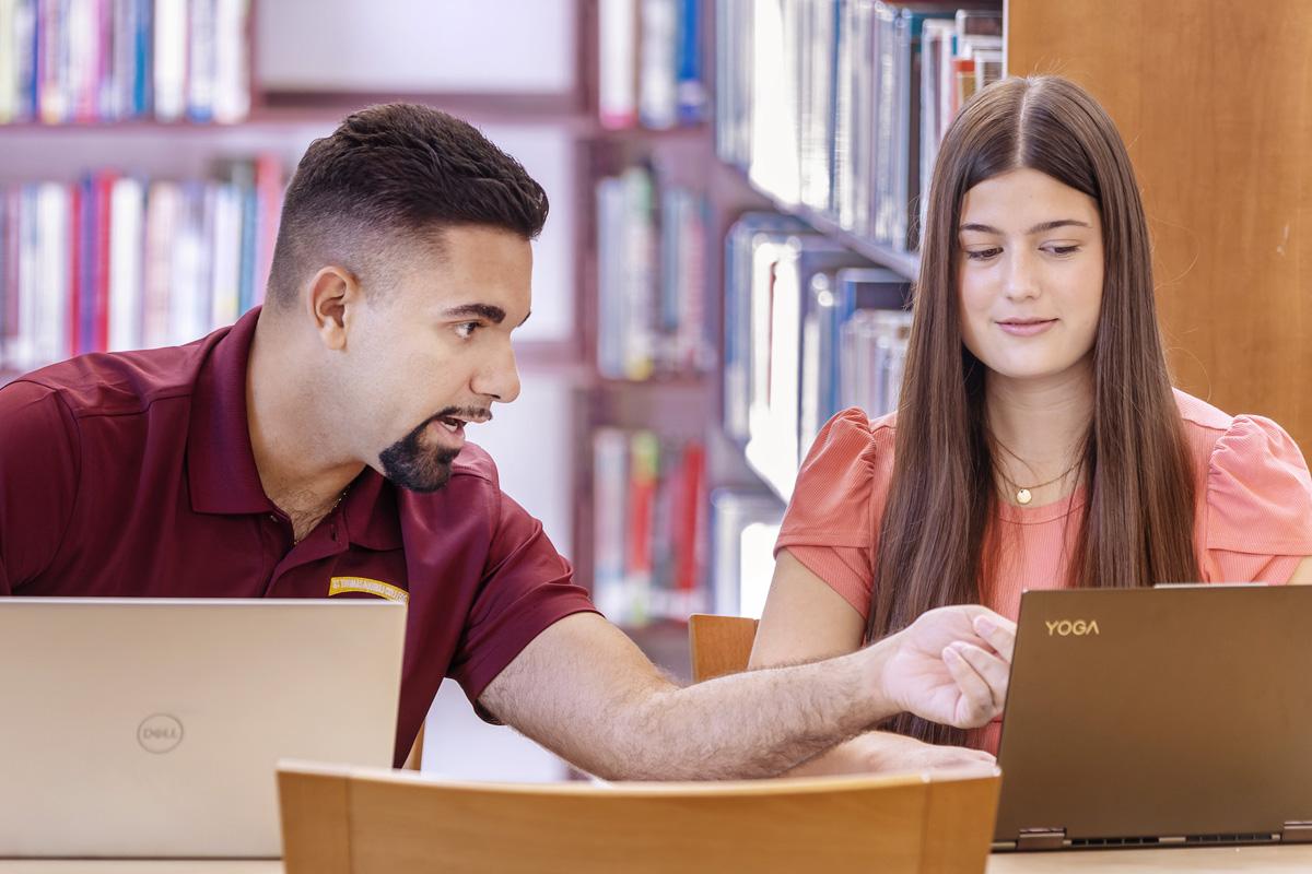 two students seated at desk in library with laptops on tables both looking at the screens one student pointing to screen
