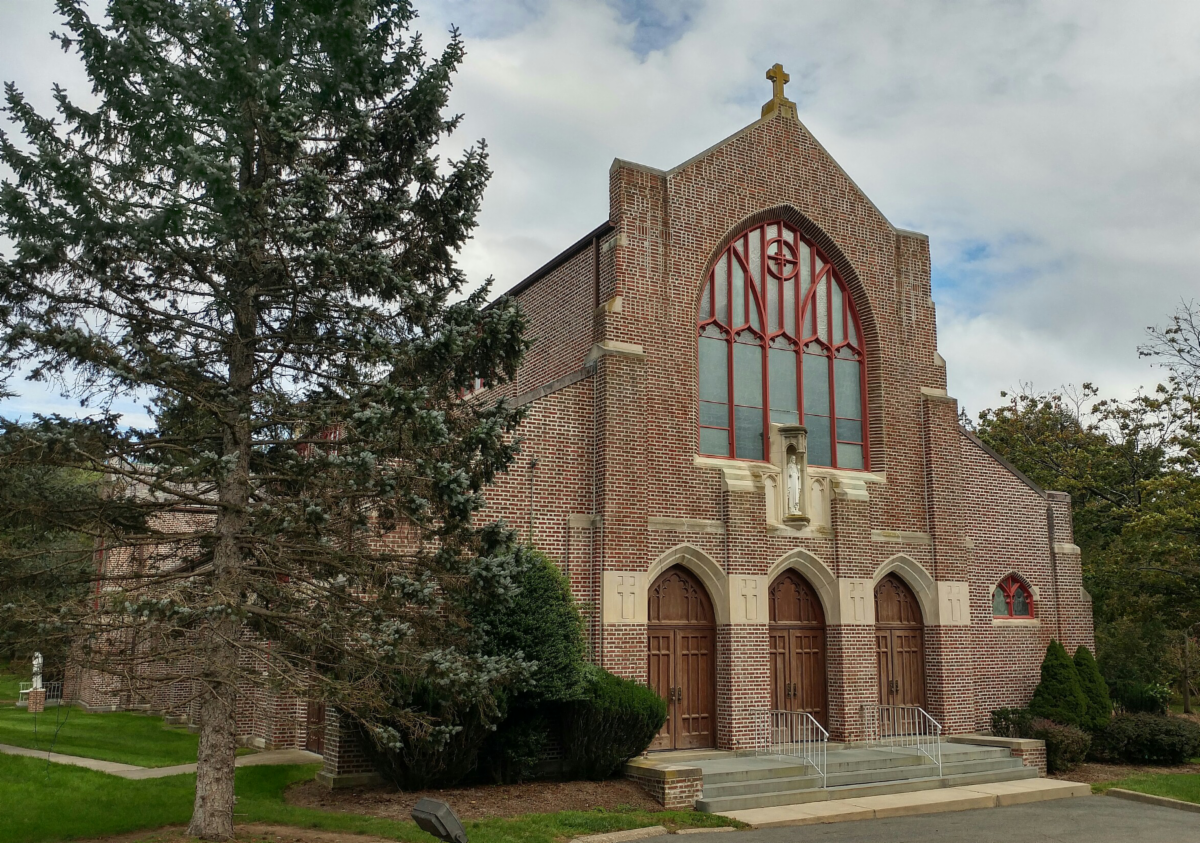 scared heart chapel brick building with cross on top three wooden doors and large window above with pine tree on left and grass and cloudy skies in background
