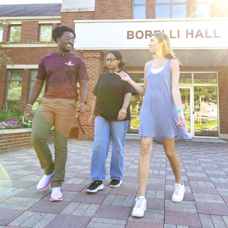 outside of borelli hall with sun shining over building and three students walking