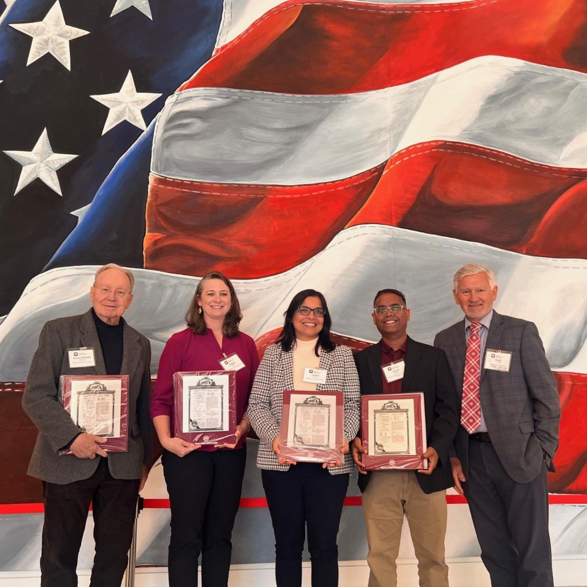 Five people standing in a row holding awards in front of an American flag. 