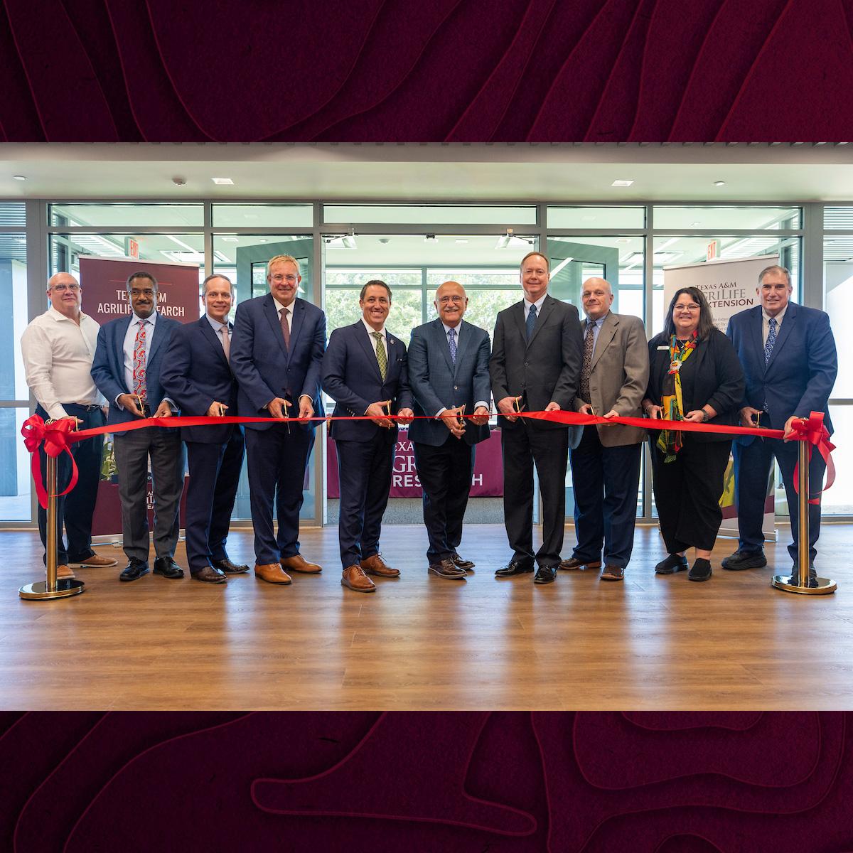 A row of people in suits indoors behind a red ribbon to be cut in ceremony