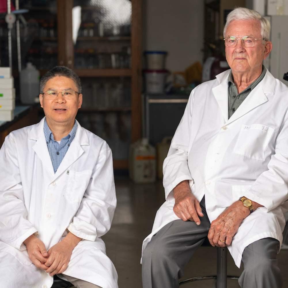 two men in lab coats sitting next to one another in a dimly lit lab