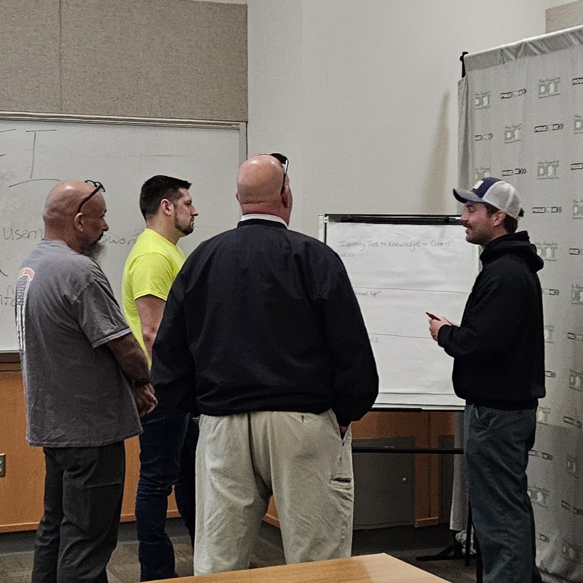 Four people stand in a small group discussion around a flip chart in a classroom or training room. One participant in a black sweatshirt and baseball cap faces the group while holding a marker and presenting information written on the flip chart. A whiteboard is visible on the left, and a branded backdrop stands along the right wall. Tables and chairs are partially visible in the foreground.