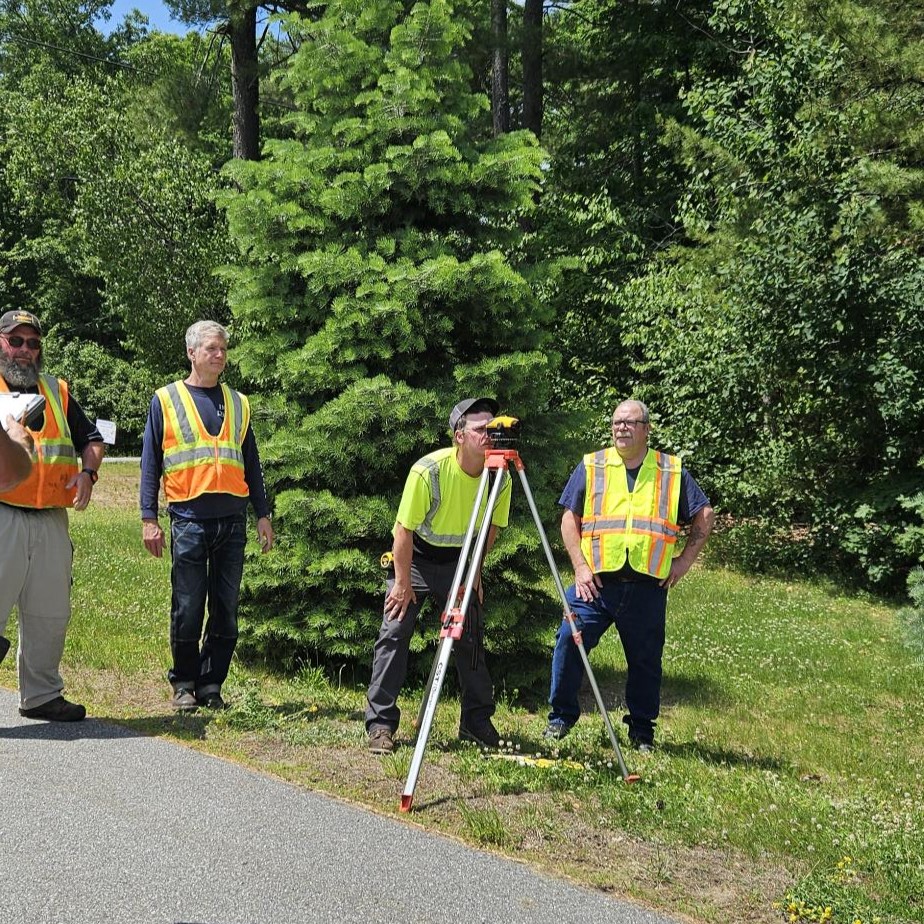 Several people wearing high-visibility safety vests stand along a rural roadside near a tractor mower and a pickup truck, surrounded by trees on a sunny day.