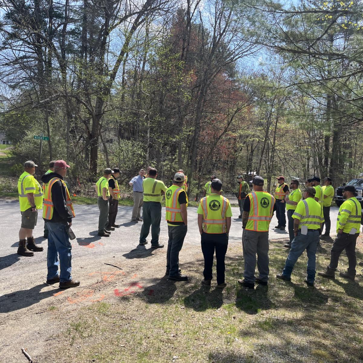 A group of approximately 15 road and utility workers wearing high-visibility safety shirts and reflective vests stand in a circle at a roadside worksite during an outdoor training session. One person in the center addresses the group while gesturing. The gathering takes place on a sunny day at a wooded intersection, with trees, utility markings on the pavement, and a street sign visible in the background.