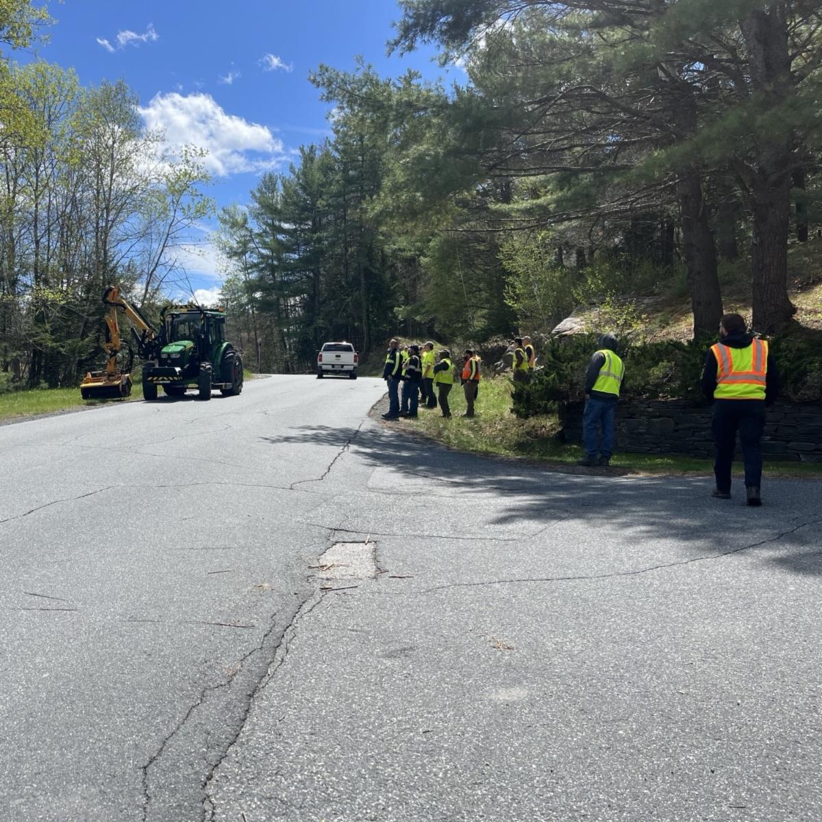 Several people wearing high-visibility safety vests stand along a rural roadside near a tractor mower and a pickup truck, surrounded by trees on a sunny day.