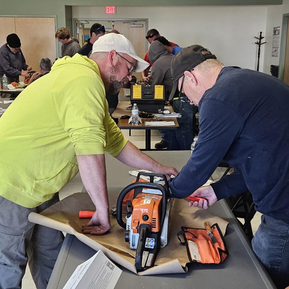 Two people work together on a chainsaw during a hands-on training session in a classroom, while several others practice with equipment at tables in the background.