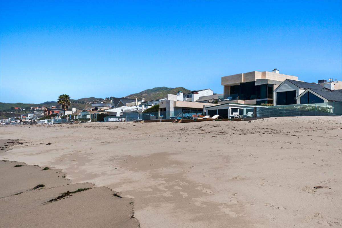 Wide beach-level view of Malibu Colony homes with sand in the foreground and coastal hills behind