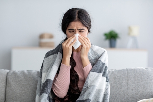 Unhappy sad young indian female in plaid suffering from fever and flu on sofa_ blowing nose in napkin in living room interior. Covid-19 lockdown_ treatment of illness_ cold and runny_ copy space