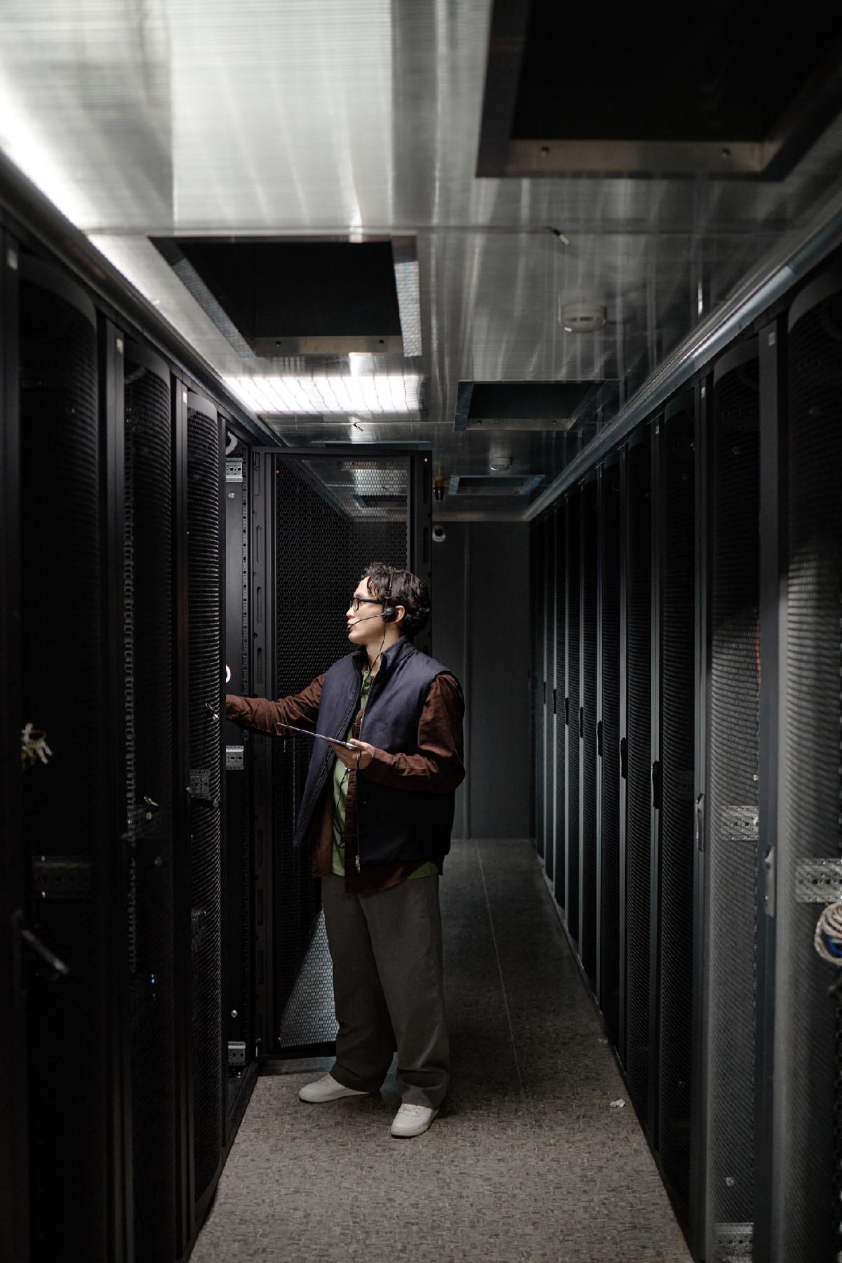 Vertical shot of Asian young adult man standing in server room inspecting server racks holding digital tablet in left hand focusing on equipment maintenance in modern data center environment