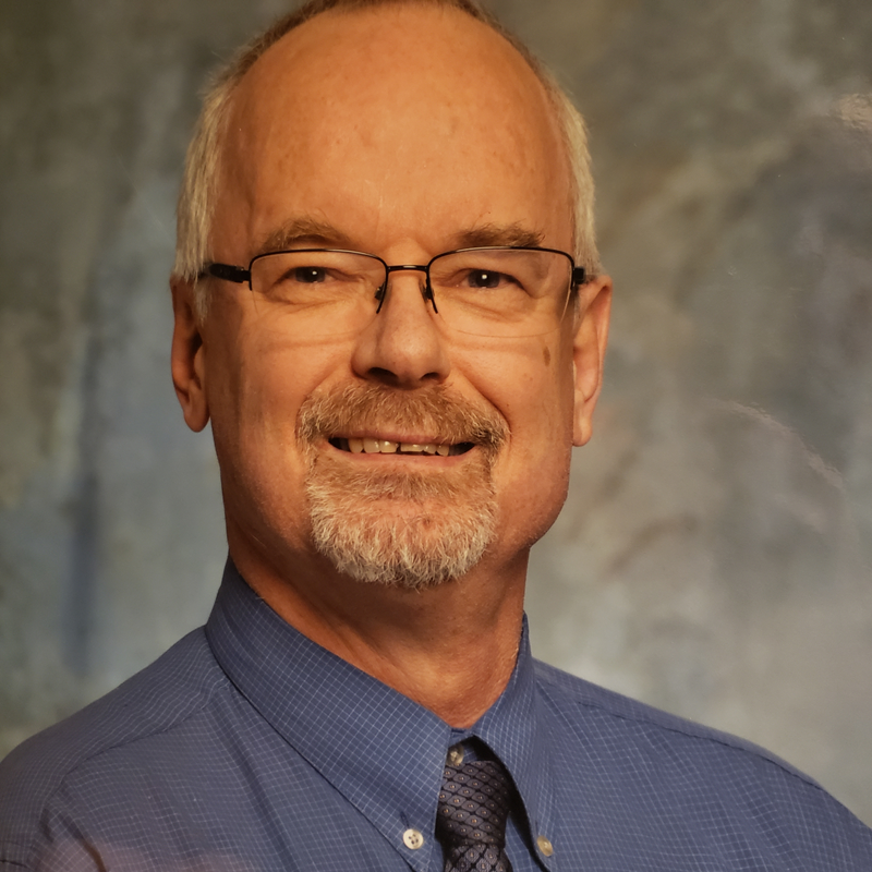 head shot of Jim Ferguson in a matte blue shirt and dark colored tie he is wearing glasses and a friendly smile
