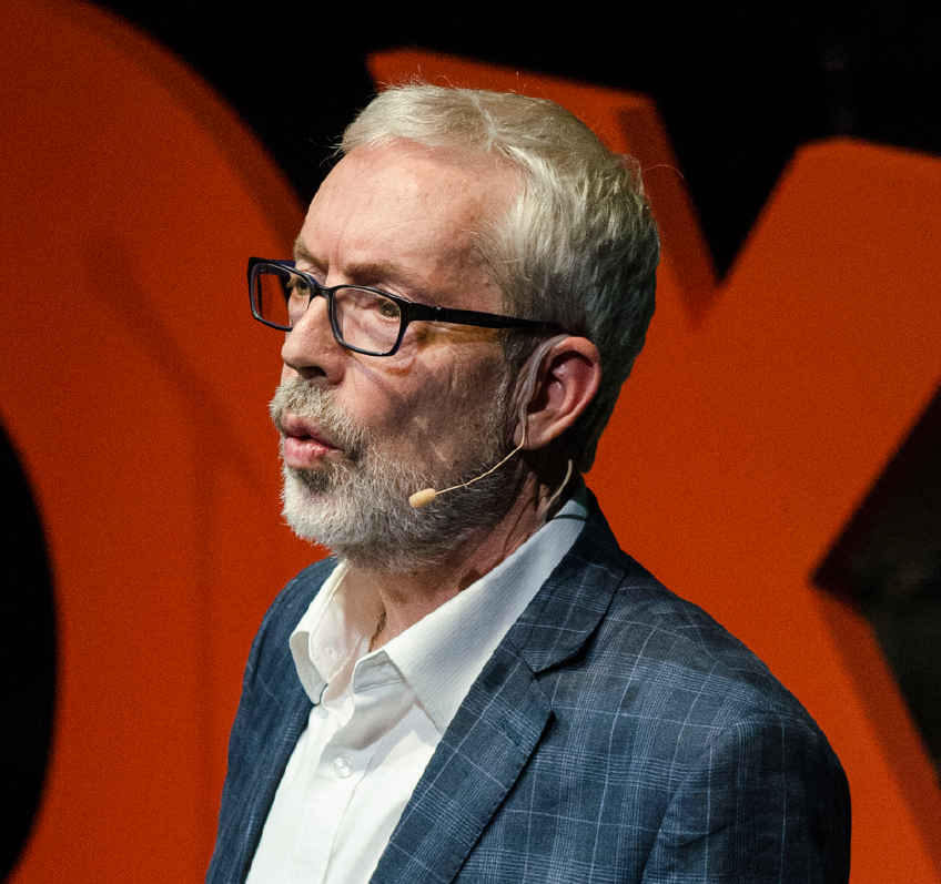 head shot of Paul Hanley in a grey plaid jacket wearing a face microphone with a red TedX sign in the background