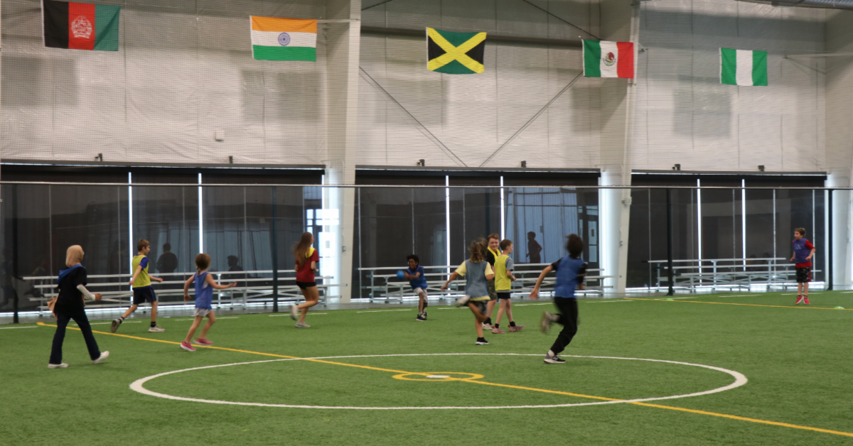 A group of youth soccer players playing at the fieldhouse, with a variety of country flags hung up including Mexico, India and Jamaica