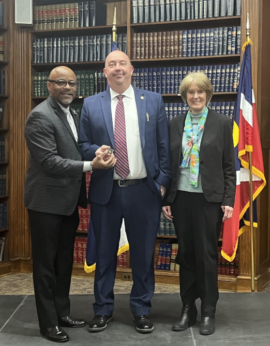 Cheif Deputy stands between Mayor Michael Hancock and DA Beth McCann in Office setting as he and the mayor hold the pin