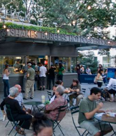 A busy cafe with outdoor seating. The cafe's sign says that it sells shakes, burgers, hot dogs, fries and sundaes.