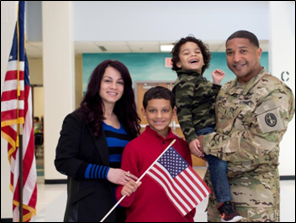Photo of a military family. The father, in uniform, stands at the right, holding a small child. At left is a mother and another son, holding an American flag.