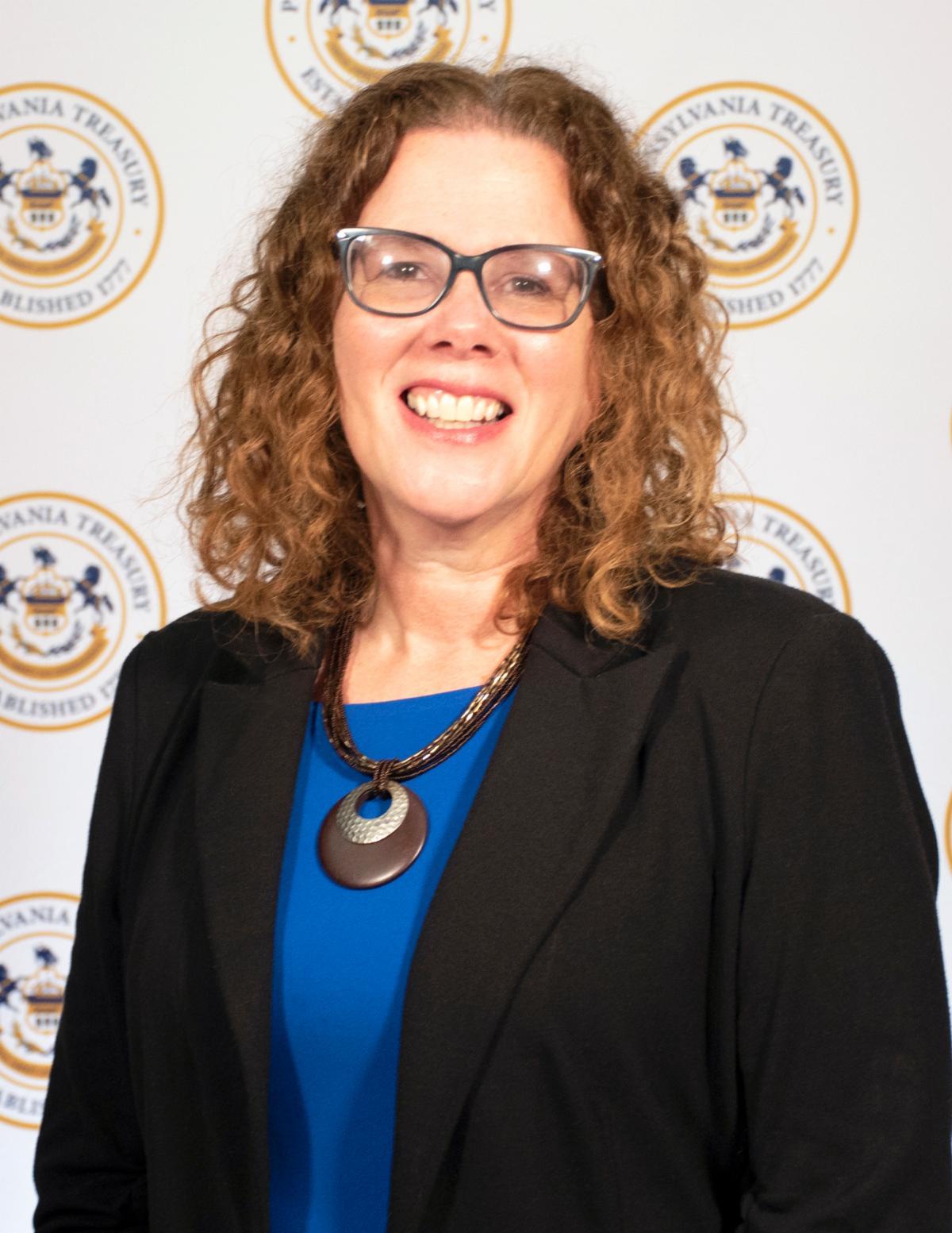 A white woman smiles, wearing a blue shirt and black suit jacket with glasses on her face.