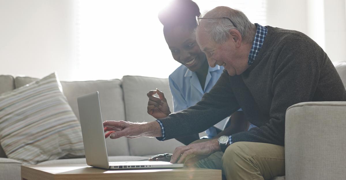 An older man and a young woman lean over a laptop on a coffee table.