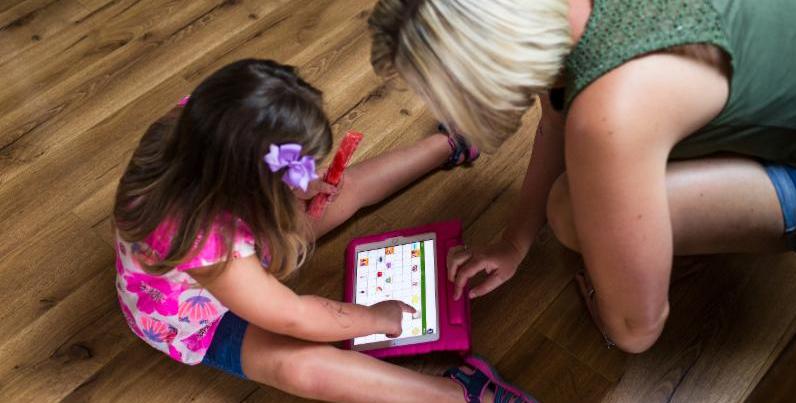 Young girl sits on the floor holding a popsicle in one hand and using the other hand to tap a communication app on a tablet screen. Her mother leans over_ also pointing to the tablet screen.
