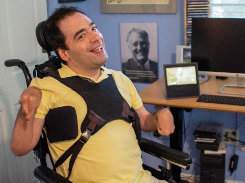 A man in a yellow polo short smiles, sitting in a power wheelchair next to a desk with a smart display on it.