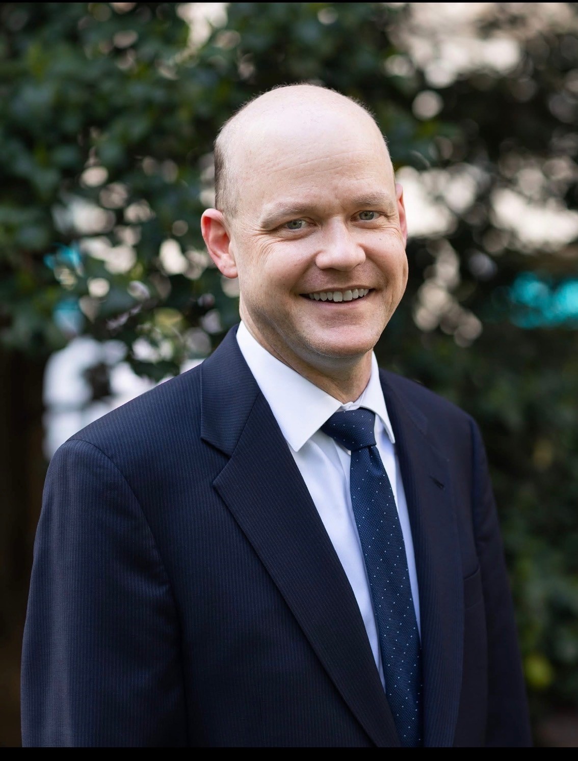 Chris is smiling and wearing a black suit jacket over a white collared shirt and tie.