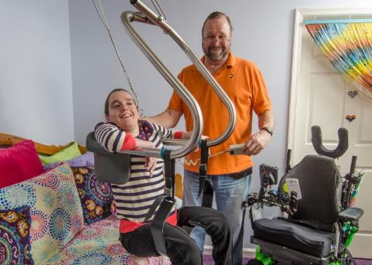 A young woman sits in a ceiling lift chair in a colorful bedroom with her father standing behind her holding the control for the lift.