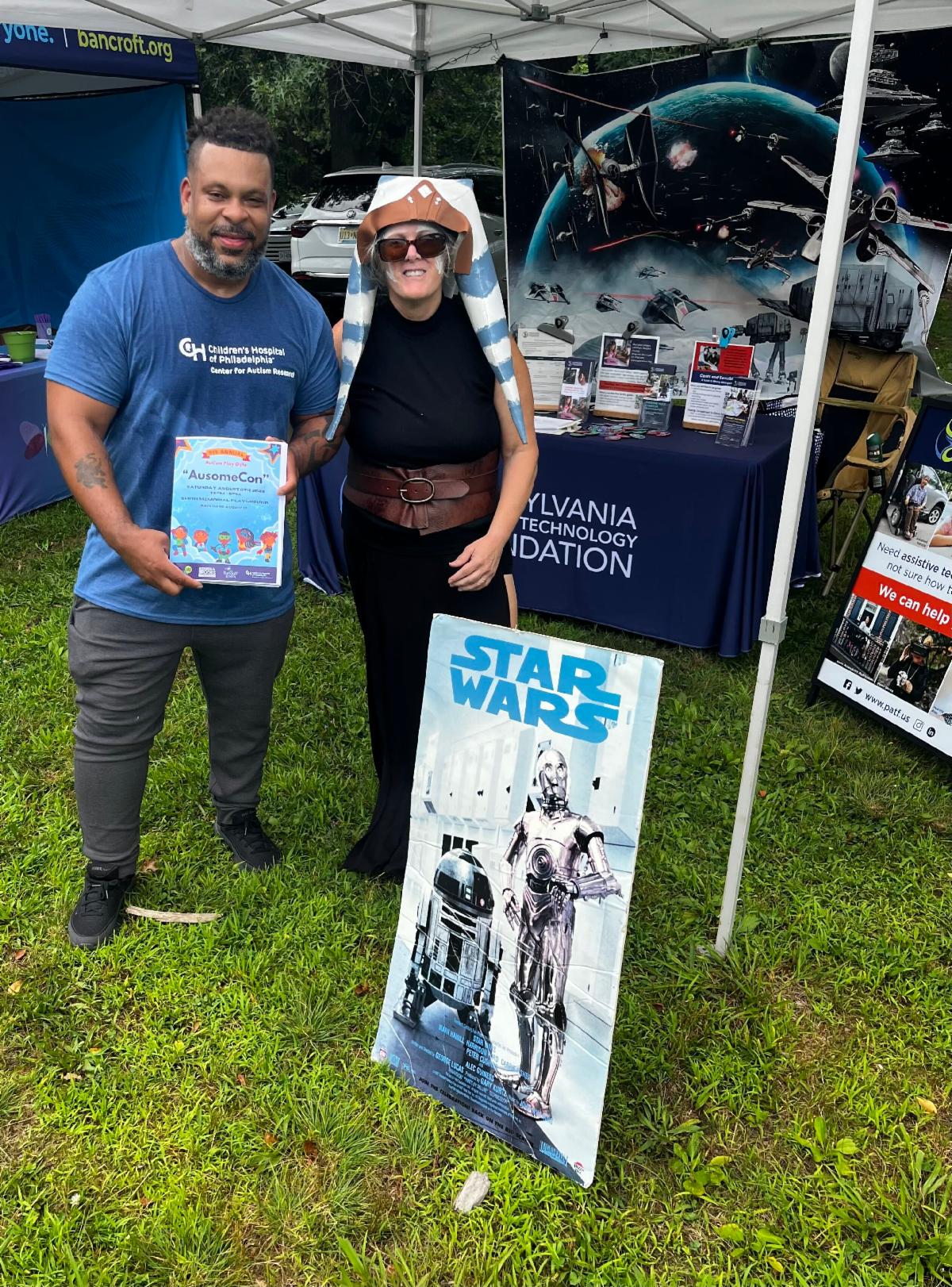 Wendy is dressed up as a character from Star Wars. She is standing next to a man in a blue CHOP shirt. They are standing behind a Star Wars poster board.