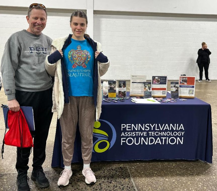 There are two people smiling together in front of PATF's resource table. The woman is showing off her shirt that says, "Kindness is always beautiful."