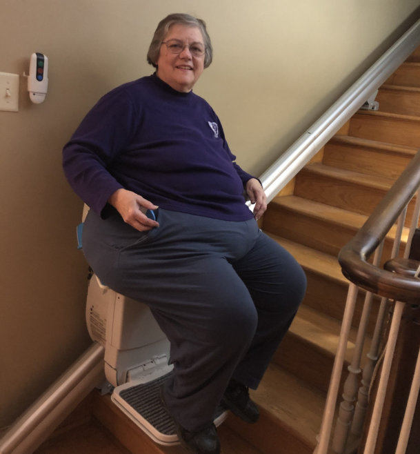 Cara Beth is sitting on her stairlift. She is wearing a long sleeve blue shirt and gray pants.
